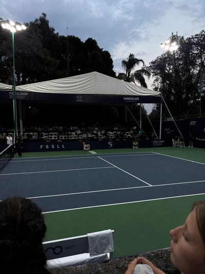 The image depicts an outdoor tennis court setup in Tetela Del Monte, Mexico, under an overcast sky, with artificial lighting activated, suggesting dusk or nighttime conditions.
In the immediate foreground, the top of a dark-haired individual's head is visible on the left, while the profile of a young female with light brown hair, dressed in light-colored clothing, is visible on the right. A light gray towel hangs over a white advertising board displaying "MORELOS OPEN" and a circular logo, situated on a textured concrete barrier that separates the viewer from the court.
The midground features a standard hard-surface tennis court. The main playing area is dark blue, bordered by a lighter green perimeter. White lines mark the court boundaries, including service lines and a baseline. A tennis net stretches across the court on the left side. No players are present on the court. A white umpire's chair is positioned to the left of the court's center, near the net. Another white chair is visible further back on the left side, and two white chairs are placed on the far right baseline.
In the background, a large white tent structure with a sloped roof provides cover for spectator seating. Beneath the tent, multiple rows of individuals are seated in white chairs, observing the court. A dark blue banner runs along the front of the spectator area, displaying sponsor logos and text including "FIDECOMP," "Mazda," "MORELOS La experiencia de México," and "FSSLL." Tall poles with bright floodlights illuminate the court; one is prominent on the left, another on the right. Dense dark green foliage and trees, some with visible purple blossoms, form a natural backdrop behind the tent and lighting structures. A single palm tree is visible on the upper right. The sky above is largely covered by gray clouds.