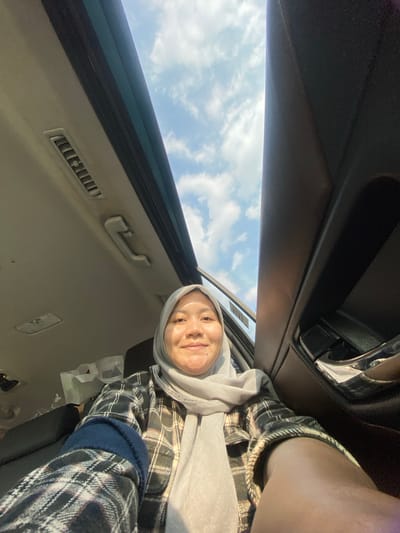 In Mandar Sari, Indonesia, a woman wearing a light grey hijab and a plaid shirt is captured in a selfie taken from inside a vehicle. The sky, with scattered clouds, is visible through the open sunroof. The interior of the car, including a grab handle and air vents on the ceiling, is also visible. The woman is smiling at the camera.