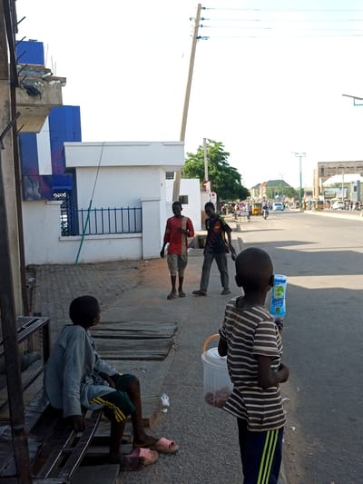 In Arawa, Nigeria, a sunny afternoon unfolds on a street. The scene features several young boys, some appearing to be street vendors. In the foreground, a boy in a striped shirt holds a plastic bottle of "Aqua Rapha" water, and a clear plastic bucket containing what looks like snacks. To his left, another boy sits on a metal bench, dressed in a grey long-sleeved shirt and shorts, wearing pink flip-flops. Further down the sidewalk, two older boys stand near a white building with blue accents. One wears a red and orange shirt and light-colored pants, while the other has on a black shirt with colorful accents and dark pants.
The background depicts a busy street with vehicles, including cars and what appear to be tuk-tuks, suggesting a bustling urban environment. A large tree and utility poles are visible, along with buildings that hint at commercial activity. The clear sky and strong shadows indicate it is likely midday. The overall atmosphere appears to be one of everyday life in a Nigerian town, with children engaged in various activities, possibly including work or play.