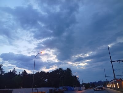 The image depicts a cloudy sky over Langenthal, Switzerland, likely during twilight. In the foreground, a road curves to the right, with a dark-colored car visible on the left side of the road. To the right of the road, there is a train, its lights illuminated, and a railway crossing signal with a barrier. Further to the right, a series of vertical lights are visible, possibly part of the train or station infrastructure. The middle ground features a line of dark trees, with a street lamp casting a warm glow on the left side of the image. In the far background, the landscape is obscured by trees and the darkening sky. Construction materials and barriers are present on the left side of the road, suggesting ongoing work.