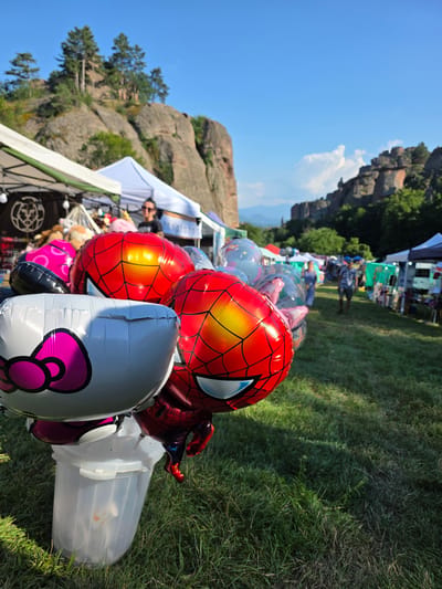 The image depicts a sunny outdoor event, likely a fair or festival, set against a backdrop of dramatic rock formations and lush green grass. In the foreground, a collection of colorful balloons, including prominent Spiderman and Hello Kitty designs, are gathered. One balloon, shaped like Spiderman, is particularly detailed with its iconic red and blue costume and web pattern. A large, clear plastic container is positioned next to the balloons, suggesting they might be for sale or distribution.
In the mid-ground, several white tents are set up, indicating stalls or booths at the event. People are visible in the background, some walking among the tents and others appearing to be tending to the stalls. The overall atmosphere suggests a lively and pleasant day.
The rock formations in the background, a key geographical feature of Belogradchik, are bathed in sunlight, hinting at a midday or early afternoon time. The clear blue sky further reinforces the impression of good weather. The presence of these unique geological features strongly suggests the location is Belogradchik, Bulgaria.
There is some visible text on a white banner behind the balloons, but it is too blurred to be legible. The image captures a moment of joyful outdoor activity, with the vibrant balloons adding a festive touch to the natural grandeur of the landscape.