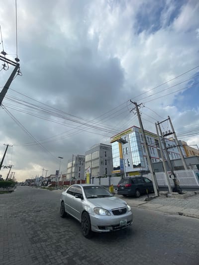 The image shows a street scene in Moba, Nigeria. The focus is on a silver car parked on a brick-paved road. A dark-colored minivan is parked behind the silver car. Several buildings are visible along the street, including a multi-story building with reflective windows and what appears to be commercial signage. Utility poles and overhead power lines are prominent in the scene, crisscrossing the sky. The sky is overcast with varying cloud cover. Other cars are visible in the distance, indicating moderate traffic. No people are clearly visible. The environment is urban.
