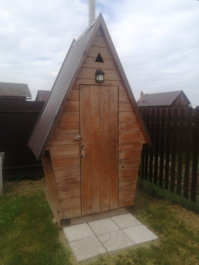 A small, wooden, A-frame shed with a metal roof stands on a patch of grass in Yemelyanovo, Russia. The shed has a single wooden door with a metal handle and a lock. Above the door, there is a small triangular vent and a wall-mounted light fixture. The shed is situated next to a dark brown wooden fence. In the background, other wooden structures and roofs are visible. The sky is overcast with some clouds. The ground in front of the shed is covered with grass and a few light-colored concrete pavers.