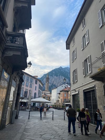 The image captures a street scene in Lecco, Italy, presenting an unusual perspective tilted to the right. Buildings line both sides of the street, showcasing a variety of architectural styles, from ornate facades to simpler, shuttered windows. A mountain rises in the distance, visible between the buildings, suggesting the proximity of the Italian Alps.
People are walking along the right side of the street, indicating pedestrian activity. A cafe or restaurant with outdoor seating is visible, suggesting a place for leisure and gathering. A KIKO Milano store front can also be seen. The street itself is paved with light-colored stone. The sky is partly cloudy, and the lighting suggests it is daytime. A clock tower can be seen in the distance.