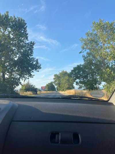 The image is a forward-facing view from inside a vehicle, looking out through the windshield onto a rural road in Graf Ignatievo, Bulgaria. The road is paved and appears to be two lanes, bordered by dry, yellow grass and tall, leafy green trees on both sides. The sky is a clear, bright blue with a few wispy white clouds.
In the distance, a large billboard is visible on the right side of the road. It is orange and white with text and images, though not clearly decipherable. The dashboard of the vehicle is in the foreground, dark gray and textured, with visible vents and controls. The overall impression is of a sunny day on a scenic drive through the Bulgarian countryside.