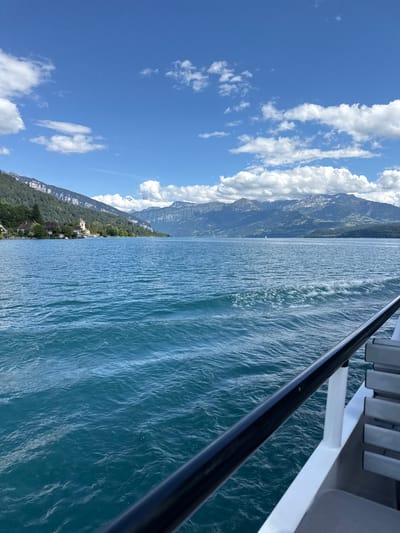 A panoramic view captures the expansive, deep blue waters of Lake Thun on a clear day, with a white boat railing and seating visible in the foreground, suggesting a passenger vessel. The water is rippled, indicating movement, and a wake trails behind the boat. In the distance, the shore is lined with lush green trees and buildings, including a distinctive castle-like structure, set against the backdrop of dramatic, layered mountains. The sky is a vibrant blue, dotted with fluffy white clouds. The scene is set in Hünibach, Switzerland, known for its scenic lake and mountain vistas.