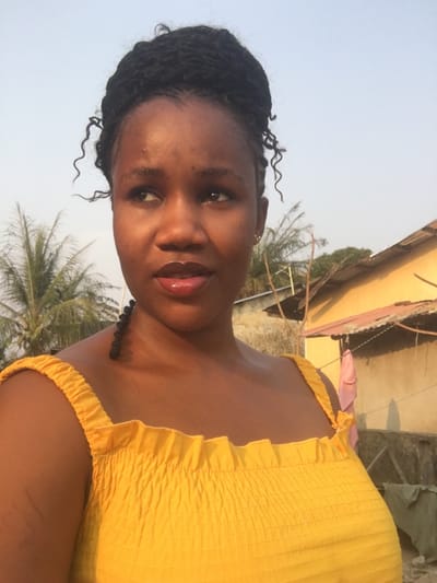The image depicts a young woman outdoors in Abagbo, Nigeria, positioned in the foreground against an urban or semi-urban backdrop. She is of African descent, with dark, braided hair styled in an updo, featuring some curly strands framing her face and a single braid resting on her right shoulder. She wears a bright yellow, smocked sleeveless top with wide shoulder straps. Her head is turned slightly to her left, and her gaze is directed upwards and to her left. Her lips appear glossy, and she has small stud earrings in both ears. In the background, under a clear, light blue sky, several green palm trees are visible. To the right, a section of a yellow-painted building with a dark corrugated metal roof is present, alongside another lighter-colored building behind it. A light pink garment is hanging on a line or structure near the yellow building.