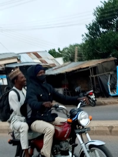 Two individuals are riding a motorcycle on a paved road in Garko, Nigeria. The person in the front is wearing a blue head covering, a dark jacket with a hood, and a dark face covering. The person in the back is wearing a light-colored traditional cap and a white shirt with a black backpack. The motorcycle is red with a silver front fender and chrome handlebars. In the background, there are several small buildings with corrugated metal roofs and some trees. The sky is overcast.