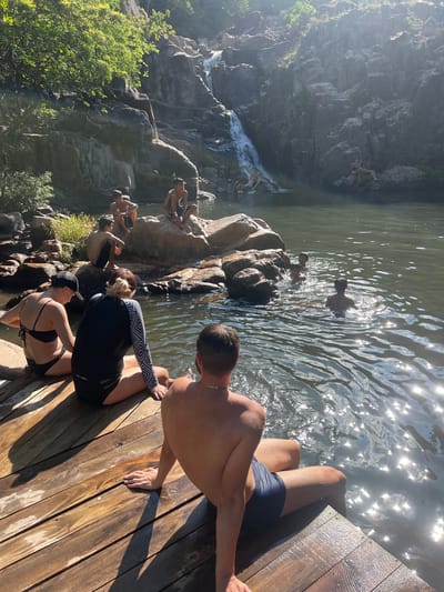 The image depicts a group of people at a natural waterfall and pool area, likely in Vĩnh Hải, Vietnam. In the foreground, two individuals are seated on a wooden platform, facing away from the camera and towards the water. To the left, a person in a black bikini and a black baseball cap is seated. Next to them, a person wearing a black long-sleeved rash guard with a geometric pattern and black bottoms is also seated. Further to the right, another individual with their back to the camera is seated on the wooden platform, wearing navy blue swim trunks.
In the water, several people are visible. Some are swimming or wading in the pool, while others are sitting on rocks near the edge of the pool or at the base of the waterfall. The waterfall cascades down a rocky cliff face, surrounded by lush green vegetation at the top. The water is relatively still, with sunlight reflecting off its surface, creating a shimmering effect. The environment appears to be a natural, outdoor setting with rock formations, trees, and a body of water. The overall atmosphere suggests a leisure activity, possibly swimming or relaxing in a natural landscape.