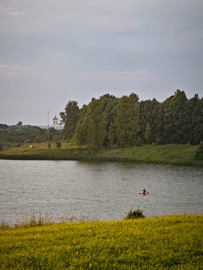 The image shows a wide, calm body of water with ripples on its surface. In the distance, a person is seen on a paddleboard in the water, facing away from the camera. The shoreline is lined with tall green grass and some sparse vegetation. Behind the shoreline, a dense line of trees forms the middle ground of the image. Further in the background, rolling hills are covered with a mix of trees and grass, and a white building with a dark spire and a cross on top is visible, likely a church. A tall, slender antenna structure is situated behind the church. The sky is overcast with a light, hazy appearance. The overall scene depicts a tranquil natural landscape.