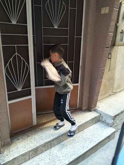 A young boy stands on concrete steps at the entrance of a building in قرية الضبية, Syria. He is seen in profile, facing left, wearing a two-toned olive green and dark gray jacket, black athletic pants with white stripes, white socks, and dark flip-flops. He holds a bundled, light-colored object, possibly a plastic bag containing fabric or goods, in his right arm. The entrance features a double metal door with decorative white grilles in a fan-like pattern, which appears partially open. To the right of the door, a textured light brown wall displays two electrical outlets or switches, a grey metal electrical box, and a large black numeral '9' painted below it. The steps are light-colored terrazzo.