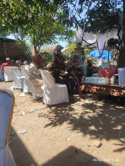 The image captures an outdoor gathering, likely a social event or celebration, under the dappled shade of trees. The scene is set on a dirt ground, with strong sunlight and shadows indicating a bright, clear day in the mid-afternoon.

In the foreground and midground, several individuals, predominantly women, are seated on white chairs, some of which are covered with white fabric and have gold trim. Most of the women are wearing headscarves (hijabs) and modest clothing, suggesting a cultural or religious context, possibly Muslim. Three women are prominent in the midground, actively eating from plates held in their laps. One woman on the left wears a light gold or beige outfit, while next to her, another woman is dressed in a dark green or brown attire. To her right, a third woman is wearing a grey outfit with striped details. They all appear relaxed, focused on their meal.

In the background, more guests are visible, some seated. A man in a red shirt and a white cap (kufi or songkok) can be seen on the far left. Further back, a woman in a light pink or purple hijab stands partially obscured.

To the right side of the image, a beautifully carved wooden table holds several clear drinking glasses, a transparent plastic jug or dispenser, and some red plastic bags or containers, suggesting a drink station or serving area. Underneath the table and around the chairs, some litter such as plastic bottles and wrappers are visible on the ground.

The setting includes decorative elements in the background, such as white and black fabric draping adorned with white floral accents, possibly indicating a temporary canopy or a decorated area for the event. A blue tarpaulin or tent structure is also visible behind these decorations. Residential buildings with light-colored walls and tiled roofs are in the distant background, suggesting a community or home-based event.

The time and date "05/02/2026 14:24" are visible in the bottom right corner of the image, confirming the photo was taken at 2:24 PM on May 2nd, 2026. The overall atmosphere appears convivial and relaxed, as people enjoy food and company outdoors.