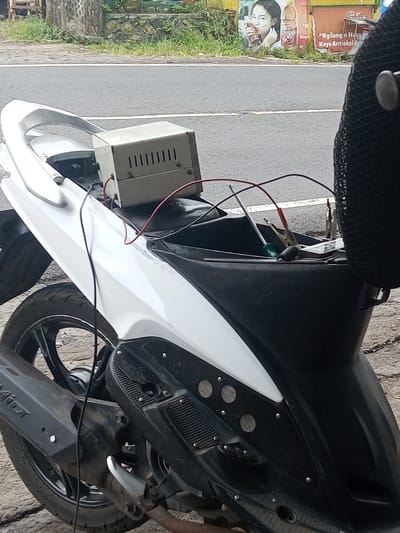 A white and black scooter is parked. Its seat is open, revealing a metal box with ventilation slots, connected with red and black wires. Tools, including a screwdriver and clips, are visible within the seat compartment. The scooter has a black exhaust and tire. In the background, there's a road, some greenery, and an advertisement featuring a woman holding a beverage.