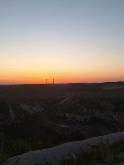 The image shows a landscape scene at sunset. The sky is a gradient of colors, transitioning from pale blue to a vibrant orange and yellow near the horizon. Two wind turbines are visible in the distance, silhouetted against the colorful sky. The foreground on the right is a rocky, uneven terrain with some vegetation. On the left side of the image, the landscape appears flatter and covered with fields. The overall mood of the image is peaceful and serene, characteristic of a sunset. The time of day is clearly sunset. No people or text are visible in the image.