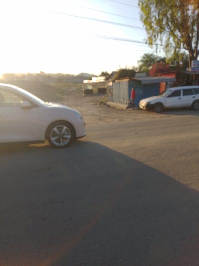 The image shows a street scene in Athi River, Kenya, taken from the perspective of a white car stopped on the road. The image is blurry and overexposed, likely due to glare from the sun. The car is facing towards the right of the frame. In the background, there is a small, blue building on the right. A person wearing a red shirt is walking toward the camera on the right. A white car is parked to the right of the person. The road is paved and there are some buildings visible in the distance. The scene is sunny and the light is bright. There are a few power lines running across the sky. The scene is likely taken during the late afternoon or early evening.