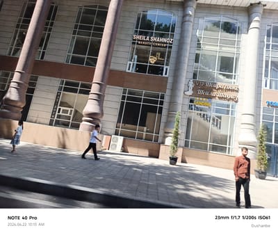 The image depicts a street scene in Dushanbe, Tajikistan, featuring a modern building with large windows and ornate columns. Two individuals are visible on the paved sidewalk. On the left, a young girl wearing a white top and a checkered skirt walks towards the right. On the right, a young man in a white t-shirt and black pants walks towards the left. Further to the right, another young man in a brown long-sleeved shirt and black pants stands facing left. Signs are visible on the building's facade, including "SHEILA SHAHNOZ BEAUTY CENTER" and other text in what appears to be Cyrillic and possibly Persian script. An outdoor air conditioning unit is visible near the building, and potted evergreen trees are placed along the sidewalk. The scene is illuminated by sunlight, casting shadows on the ground. The bottom left of the image shows a timestamp "2026.06.22 10:13 AM" and the device "NOTE 40 Pro". The bottom right corner indicates the location as "Dushanbe" and camera settings "23mm f/1.7 1/200s ISO61".