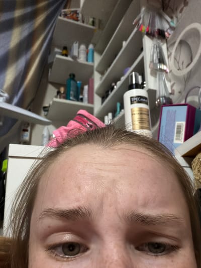 A close-up, angled image shows the face of a person with light skin, visible eyebrows, and eyes. The background features a shelving unit filled with various items, including containers and bottles of beauty products, nail polish, and some boxes. The overall scene appears to be in a private indoor space, likely a bedroom or bathroom. The image does not contain sufficient information to determine if it was taken in Berezniki, Russia or any other specific location.