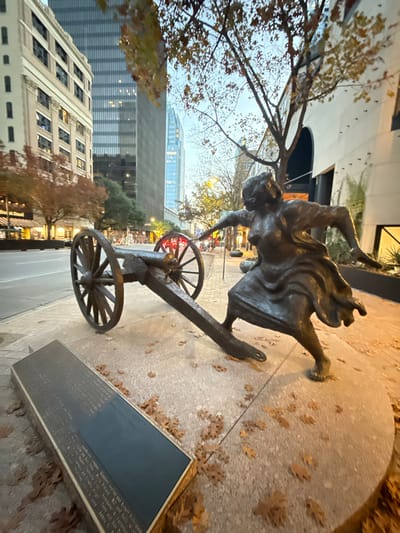 A bronze statue depicts a woman urgently pulling a small cannon, her dress flowing. The statue is positioned on a sidewalk scattered with fallen leaves. In the background, a mix of tall buildings rises, with the sky visible above. A plaque is affixed to the ground nearby, presumably providing information about the statue or its historical context. The statue and surroundings suggest a public art installation in an urban environment, likely commemorating an event or figure.