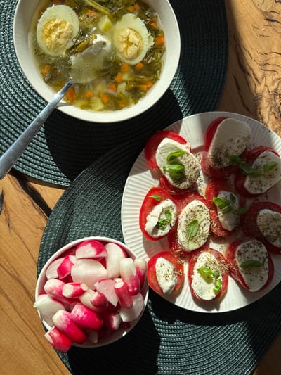 This top-down, natural light photograph captures a rustic table setting featuring a meal in what appears to be a home kitchen in Steklyannyi, Russia. The scene is set on a wooden table, with a dark, woven placemat underneath two dishes and a bowl.
The primary subjects are the food items. A white plate holds a colorful caprese salad, composed of alternating slices of ripe red tomatoes and fresh white mozzarella, each topped with a sprig of basil and a sprinkle of dried herbs. Adjacent to the caprese salad is a white bowl filled with sliced radishes, their vibrant pink and white hues contrasting with the dark placemat. To the left, a white bowl contains a hearty vegetable soup. The soup is a clear, green broth filled with leafy greens, diced carrots, potatoes, and two halves of a hard-boiled egg. A sliver of sour cream sits on top of the soup, and a silver spoon rests on the edge of the bowl.
The natural light suggests it is daytime, likely late morning or early afternoon, given the warm shadows cast across the table. The overall impression is one of a simple, healthy, and home-cooked meal. There is no visible text in the image.