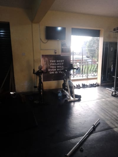 The image depicts the interior of a fitness facility located in Njoro ward, Kenya, characterized by light yellow walls and a dark, matted floor. The room contains two stationary exercise bikes: one dark-colored positioned centrally and another lighter-colored bike situated to its right. A blue plastic water bottle rests on the floor between the two bikes. Mounted on the left wall is a television screen, below which a large banner displays the motivational text: "THE BEST PROJECT YOU WILL EVER WORK ON IS YOU." A smaller white sign, likely containing gym information, is affixed to the wall near the television. To the far left, several vertically stacked dark objects, possibly mats or panels, are visible. The right side of the room features a large mirror reflecting the interior.
An open doorway or balcony area dominates the right-center, allowing significant natural light to enter and illuminating a row of approximately ten pairs of assorted athletic shoes neatly lined up on the floor. Beyond the doorway, a metal railing, green foliage, and distant structures are discernible under a bright sky. On the dark floor in the foreground, a silver barbell, devoid of weights, lies horizontally, accompanied by a single circular weight collar. No individuals are present in the scene, and no active interactions or movements are depicted. Electrical conduits are visible running along the walls.