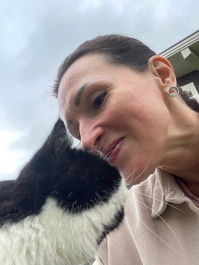 A close-up, low-angle, outdoor, selfie-style photo shows a woman from the chin up, cuddling with a black and white cat. The woman has dark brown hair, dark eyebrows, and appears to be smiling gently. She is wearing a light pink corduroy jacket and a silver hoop earring. The cat, partially obscuring the woman's face, has black fur on its head and white fur on its chest. The background is an overcast sky and a portion of a building with beige siding. The scene is likely located in Solov'evka, Russia, given the general style of architecture visible.