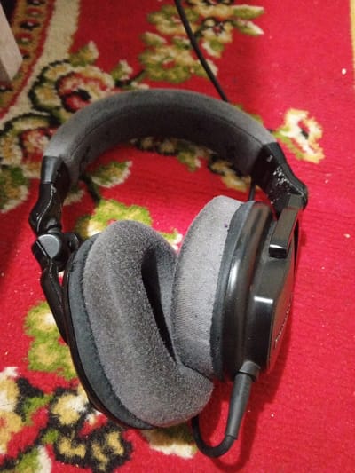 A pair of black over-ear headphones with a padded headband and gray fabric earcups rests on a red patterned carpet in Kpein, Liberia. The headphones exhibit a black plastic frame with visible wear on the joints connecting the headband to the earcups. The gray fabric ear cushions appear worn and show minor dust accumulation. A black audio cable extends from the base of the right earcup, curling towards the bottom of the frame. The carpet features a repeating floral motif with elements in yellow and green. No individuals or active interactions are present in the scene.