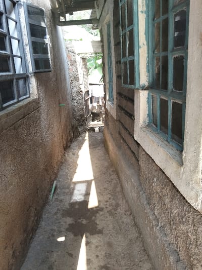The scene depicts a narrow outdoor passageway between two buildings in Nyansiongo ward, Kenya. The building on the left features a rough, earth-toned wall with two dark-framed, multi-pane windows. A green pipe segment is visible horizontally along this wall. The building on the right presents a lighter, textured wall with multiple light blue-framed, multi-pane windows, where the paint on the frames shows extensive peeling. The ground is concrete, exhibiting dark, irregular stains consistent with moisture and distinct, elongated shadows cast by direct overhead sunlight. Above the passageway, a black pipe and a lighter-colored pipe run horizontally. The far end of the passage opens to a brighter area, revealing additional structures and green foliage, including broad banana leaves. No individuals or discernible actions are present.
