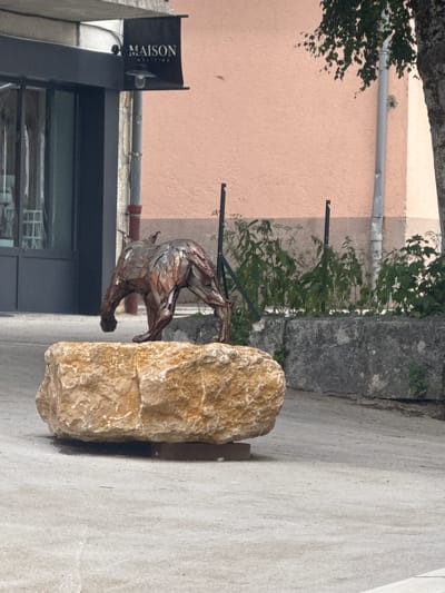 In the town of Les Rousses, France, a metal sculpture of a dog-like animal stands on a large, rough-hewn sandstone rock. The sculpture, made of riveted metal pieces, depicts the animal in a walking stance with its head lowered. Behind the sculpture, a light pink building with a grey downspout runs vertically. Above the building, a dark awning with a sign reading "MAISON MELITINE" is visible. The ground is paved with a light-colored, textured surface. To the right of the sculpture, a tall, dark tree trunk is partially visible. In the background, green foliage grows behind a low, grey stone wall.