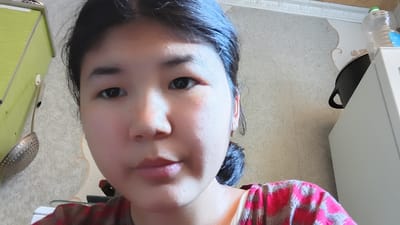 This media file is a close-up, selfie-style photograph of a young woman with dark hair pulled back in a bun. She has dark eyes, fair skin, and is looking directly at the camera with a neutral expression. She is wearing a red and gray patterned shirt.
In the background, to the left, is a portion of a lime green cabinet or drawer. A silver ladle with a perforated bowl is hanging from a hook on the cabinet. The wall behind the woman is textured and appears to be a light beige or gray color, with a decorative border at the top. To the right of the woman, a black pot with a handle is partially visible, sitting on top of a white appliance, possibly a refrigerator or a cabinet. A clear plastic bottle with a blue label is also on top of the white appliance.
The lighting suggests it is daytime, and the indoor setting appears to be a home or kitchen environment in Uchkun, Kyrgyzstan. No specific activities are depicted beyond the woman taking a photo of herself.