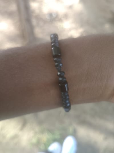 The image is a close-up of a person's arm wearing a beaded bracelet. The bracelet is made of dark, possibly black, beads, some of which are cylindrical and others round. The arm is tanned and appears to be hairless. The background is blurred and out of focus, suggesting an outdoor setting with soft lighting. The ground in the background appears to be dirt or sand, with some hints of green vegetation and lighter colored objects that might be footwear. The overall impression is a casual, everyday moment captured. There is no visible indication of the city of Majemu, Nigeria in the image itself.