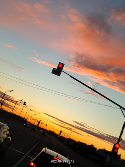 This media file captures a dramatic sunset over a highway in Ulyanino, Russia. The sky is a canvas of vibrant colors, with streaks of orange, pink, and light blue clouds against a pale blue sky. The setting sun casts a warm glow, illuminating the clouds and creating a serene yet dynamic atmosphere.
The scene depicts a busy highway with several cars on the road, suggesting ongoing traffic despite the late hour. A traffic light is visible, illuminated with a red signal, indicating that vehicles are stopping. Streetlights line the road, their warm lights beginning to punctuate the darkening sky. The perspective is from within a vehicle, looking out towards the road and the expansive sky.
Notable details include the date and time stamp "2026.06.25 21:12" overlayed on the image, placing the scene at approximately 9:12 PM on June 25, 2026. The weather appears clear with scattered clouds. The overall mood is one of calm and beauty, a fleeting moment of natural splendor observed during daily life on the road. The image evokes a sense of transience, with the vibrant sunset signifying the end of the day.