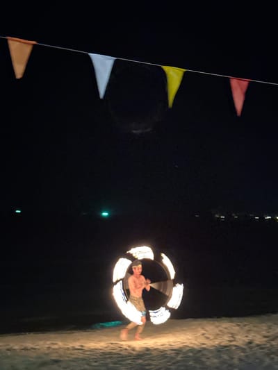 At night in Hat Khai Muk, Thailand, a shirtless man spins a circle of fire. The location appears to be a beach or sandy area. A string of triangular flags, mostly red, yellow, white, and orange, is to the left, and dim lights are in the background.