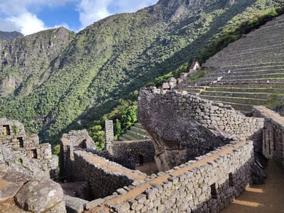 Stone ruins of Machu Picchu sprawl across the foreground with carefully constructed walls and rectangular structures. The landscape ascends into terraced hillsides, dotted with small stone buildings and active with tourists. In the background, a towering mountain densely covered with green vegetation dominates the skyline, partially shadowed by sunlight. The bright sky with scattered clouds tops the scene.