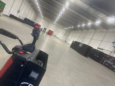 The image depicts the interior of a large, modern warehouse or distribution center in Schweitenkirchen, Germany. The space is vast, with high ceilings and a concrete floor. Rows of stacked black plastic pallets are visible in the background, extending down the length of the building. The lighting appears to be provided by long, linear LED fixtures mounted on the ceiling.
In the foreground, a red and black electric pallet jack is positioned on the left side of the frame. Its handle and control panel are clearly visible, with a red emergency stop button on the top. A compartment on the pallet jack is open, revealing some internal components, possibly a battery or control unit. The pallet jack is not in motion.
The overall environment suggests an active but currently quiet commercial or industrial setting. There are no people visible in the image. The warehouse appears to be clean and well-maintained. The pallet stacks suggest a system for storing and organizing goods.