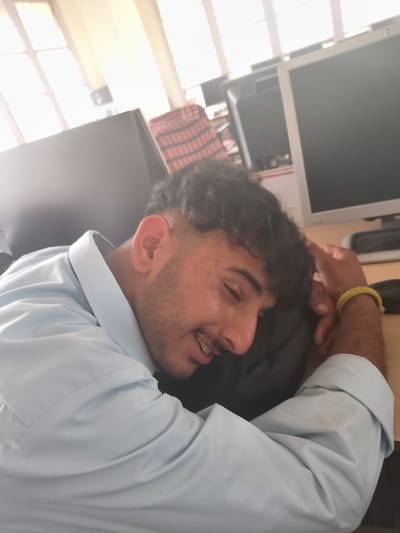 A young man with dark curly hair and a light blue collared shirt is asleep with his head resting on a dark surface. He has stubble on his chin and is wearing a yellow bracelet on his right wrist. Behind him, a computer monitor is visible on a desk. The scene appears to be an indoor office environment, possibly in Chelian, India, with a window in the background showing daylight. The image is taken from a close-up, slightly angled perspective, focusing on the man's face and upper body.
