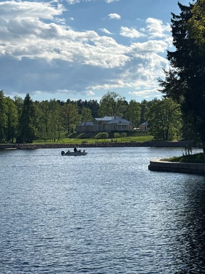 The image depicts a serene landscape with a waterway running vertically. On the left side of the waterway, there is a building nestled among lush green trees and grass. A small boat, occupied by people, is visible on the water near the shoreline. The sky above is filled with fluffy white clouds against a blue backdrop. The right side of the image is dominated by the water, and there is a stone structure along the edge of the waterway.
The location is likely within the city of Vyborg, Russia, known for its waterways and natural beauty. The building appears to be a residential or estate property, and the boat suggests recreational activity on the water. The overall impression is one of tranquility and natural harmony.