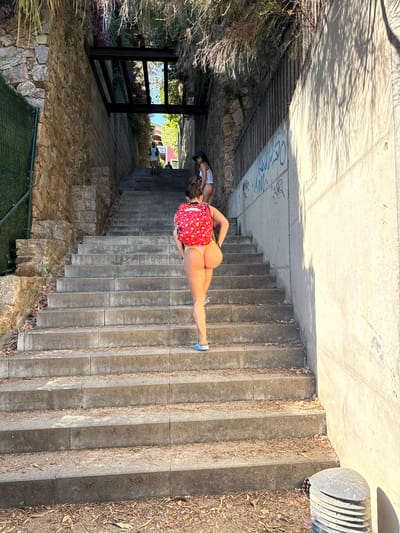 In the city of Castell d'Aro, Platja d'Aro i s'Agaró, Spain, a person with a red backpack adorned with white polka dots walks up a wide stone staircase. They are wearing a light-colored thong and blue flip-flops. The staircase is flanked by a stone wall on the left and a concrete wall with graffiti on the right. Lush vegetation hangs from above, casting shadows on the scene. In the background, other individuals can be seen ascending the stairs, and buildings are visible at the top. The ground at the base of the stairs is covered with fallen leaves and debris.