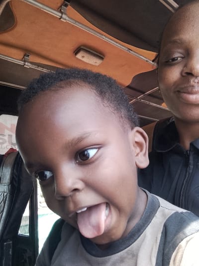 A young child sticks out their tongue with a playful expression, looking to the left. Their skin is dark, and they wear a gray and black striped shirt. Behind the child is a person with a neutral expression. The background shows the interior of a vehicle with an orange roof, metal bars, and a black seat. A small light fixture is visible on the roof.