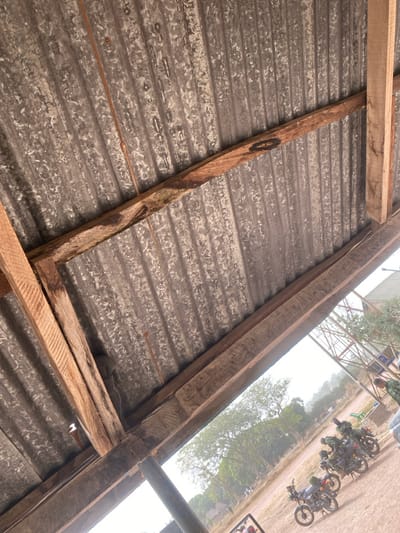 The image depicts an outdoor scene in Zaki Biyem, Nigeria, observed from under a corrugated galvanized metal roof supported by untreated wooden beams. The roof shows signs of wear, including discoloration and possible rust. A thin, reddish-brown wire or string runs along one of the corrugated metal channels. Beyond the shelter, an open area of dirt or hard-packed earth is visible under a bright, possibly hazy sky.
In the mid-ground, a minimum of three dark-colored utility motorcycles are parked in a staggered line, facing generally right-to-left. Equipment or bags are visible on some of the motorcycles. Several individuals, clad in camouflage-patterned uniforms, are present in the vicinity of the motorcycles and further into the background. One figure is partially visible near the right edge of the frame, close to a grey vehicle. Distant trees with green foliage line the horizon. A light-colored roof and a metal lattice structure are visible in the far background on the right. The overall perspective is angled upwards and tilted diagonally.