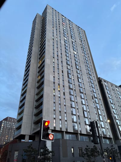 The image captures a low-angle view of a high-rise building in Manchester, United Kingdom. The building is characterized by a repetitive pattern of rectangular windows set into a gray facade. A smaller, adjacent section of the building is set back slightly, creating a tiered effect. A traffic light, displaying a red light and a "no U-turn" sign, is positioned prominently in the foreground. The sky is partially visible, with a blue hue fading towards the horizon. Lower buildings and trees are also visible.