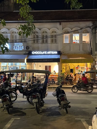 The image depicts a street scene at night in Siem Reap, Cambodia, likely along a commercial strip. The foreground is dominated by several parked motorcycles and a tuk-tuk. In the middle ground, storefronts are visible, illuminated by neon signs. One sign reads "KABAS CONCEPT STORE," another "CHOCOLATE ARTISAN," and a yellow awning displays "Angkor Hand-Pulled Noodles & Dumplings II." People are seated at tables outside some of these establishments. The background shows the upper floors of buildings with arched windows. A tree with green leaves hangs into the frame from the top left. The street is paved and appears wet, possibly from recent rain. The overall atmosphere is that of a lively evening in a tourist-oriented area.