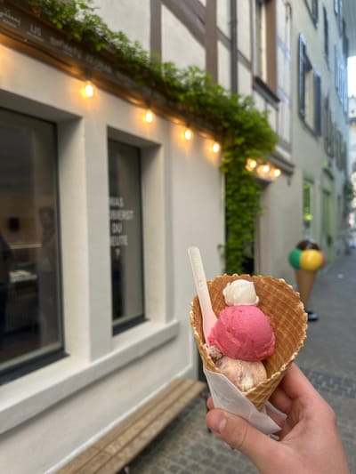 A hand holds an ice cream cone with three scoops of ice cream (white, pink, and beige) and a small white plastic spoon. The cone is wrapped in a white paper napkin. The background features a building with half-timbered architecture, illuminated by string lights. Greenery and flowers adorn the building's facade. Further in the background, another decorative ice cream cone is visible above the storefront. The storefront has windows and a sign that reads "WAS BIETEST DU HEUTE?". The location appears to be in Winterthur, Switzerland.