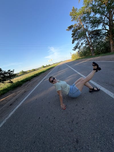 A woman is lying on an asphalt road in Foki, Russia, with her legs raised in the air and crossed at the ankles. She is wearing a horizontally striped shirt, denim shorts, and black sandals with thick soles. She has sunglasses on and is looking towards the camera. One arm is extended to her side. A solid white line runs along the road, seemingly splitting the road into two lanes. Trees and vegetation line the left side of the road, and a clear blue sky is visible above. The road surface appears to be in good condition, with no visible cracks or potholes.
