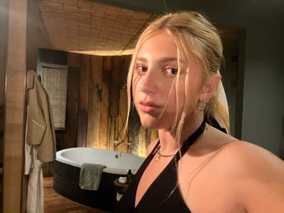 A fair-skinned young woman with blonde hair is the focal point of a selfie taken indoors, likely in a bathroom. She wears a black halter-top and gold jewelry, including hoop earrings and a necklace. Her hair is partially up, with some strands framing her face. Behind her, a freestanding bathtub with a grey towel is visible. To the left, a coat and robe hang on a wooden structure. The walls are constructed from wood planks, and a woven bamboo-like material covers the ceiling. The room has dim lighting. Given the instruction, this location is presumed to be in Livingstone, Zambia.