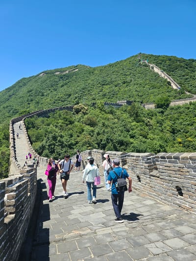 In Huairou District, China, a group of tourists is observed walking along a section of the Great Wall under a clear blue sky. The Great Wall is constructed with stone and brick, with watchtowers visible in the distance along the undulating mountainous terrain covered in lush green vegetation. Several individuals are captured in various poses of movement, some are walking forward, while others are engaged in conversation. The focus is on the diverse group of people, their clothing, and their interaction with the historical landmark. The environment is characterized by the immense scale of the Great Wall, the surrounding natural landscape, and the bright sunlight casting shadows on the paved walkway.