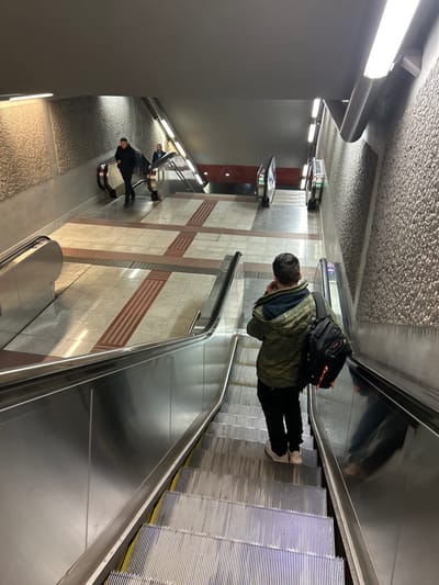 An interior view depicts a modern public transportation station, likely a metro, characterized by multiple escalators and moving walkways. The perspective is slightly elevated, looking down and across the platforms.
In the mid-ground, a person is seen from behind, traveling upwards on a metal escalator or moving walkway. They are wearing a dark olive-green jacket over a dark top, black pants, and light-colored sneakers. A black backpack with subtle orange accents is worn on their back. Their head is slightly tilted downwards, suggesting they might be looking at a device in their hands.
Further down the station, to the left of the main subject, another individual is visible, also from behind. This person wears a dark jacket and appears to be standing on another moving platform or a nearby landing.
The station's architecture includes a light grey, textured ceiling and walls tiled with a geometric pattern of light-colored and grey rectangular sections. Horizontal dark red striped patterns are integrated into the wall tiling. Bright fluorescent lighting fixtures are visible both on the ceiling and along the side walls, illuminating the entire space. Shiny silver handrails and the metallic surfaces of the escalators dominate the foreground and mid-ground. In the background, several grey pillars feature illuminated digital signs or displays, though the text on them is not legible.
The scene captures the routine activity of commuters using the modern facilities of a transit station. The clean, well-lit environment suggests a contemporary design.