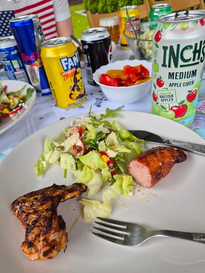 Here's a description of the image:

**Scene:** The image shows a partly eaten plate of food on a table. The food includes a sausage cut in half, a piece of grilled chicken, and a mixed salad with lettuce, radishes, corn, and other ingredients.

**Objects:**
* A white plate with food
* Fork and knife
* Cans of various drinks: Inch's cider, Fanta Pineapple, Red Bull, and others that are partially visible.
* A bowl of roasted peppers.
* A table with a tablecloth with stars on it.

**Subjects:** The focus is on the partially eaten meal. A person's arm wearing a white shirt with a part of the American flag is partially visible in the background.

**Environment:** The scene appears to be outdoors, possibly a picnic or a barbecue setting.

**Location:** The presence of Inch's Cider indicates the West Suffolk, United Kingdom.