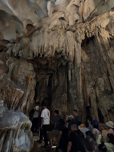 The image captures a group of people exploring a cavernous natural cave, likely in Ha Long Ward, Vietnam. The scene is dominated by intricate and varied rock formations, exhibiting a range of textures from smooth, undulating flowstone to rugged, layered, and jagged surfaces. The predominant colors are shades of gray and brown, with occasional hints of warmer, reddish-orange tones on some rock faces, suggesting mineral deposits.
A diverse group of tourists, casually dressed in t-shirts, shirts, and hats, occupies the right side of the frame. They appear to be walking along a path or elevated walkway within the cave, their bodies angled as if observing the surroundings or progressing through the space. The perspective is somewhat elevated, looking down upon the visitors and the descending path.
The lighting within the cave appears to be artificial, strategically illuminating the geological features and the path, creating contrasts between the well-lit formations and darker recesses. No specific time of day or weather can be discerned from the enclosed environment. The activity clearly depicts a guided tour or exploration of a natural cave system, a common tourist activity in the Ha Long region, renowned for its karst landscapes and grottoes. Emotions are not distinctly visible, but the people seem engaged in sightseeing. There is no clearly decipherable text visible on any objects or surfaces within the frame.