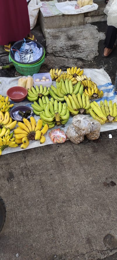 The image shows a street vendor selling bananas and other snacks in Kecamatan Lawang, Indonesia, at the coordinates -7.846 latitude and 112.7 longitude. The vendor has laid out a large white cloth on the ground, on which they have arranged bananas, eggs, snacks wrapped in plastic, and a few bowls. The bananas are arranged in groups, some ripe and yellow, some still green. The street is paved with gray concrete, with a few cracks and some small, dark patches on the surface. A partially visible tire sits on the left side of the image, with a blurry figure in the upper left. An individual's feet are visible on the right side of the image. The scene appears to be a typical street market setting in Indonesia.