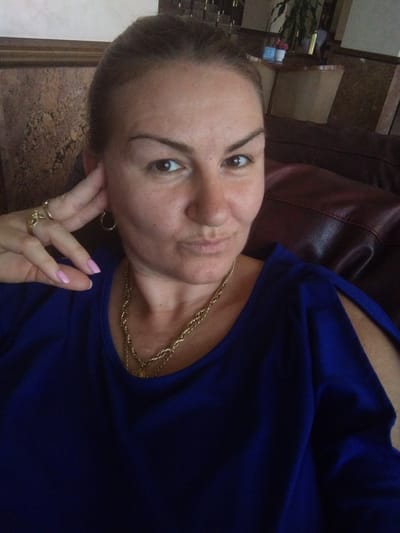 This is a close-up, vertically oriented, first-person photograph of a woman. She is wearing a royal blue top and a gold chain necklace. Her right hand is raised, with fingers adorned with two gold rings, touching her head near her ear. Her fingernails are painted a light pink. The woman's expression is relaxed. She is positioned on a dark red, possibly leather, couch. The background appears to be an indoor setting, with what looks like a textured countertop and wooden furniture visible. The lighting suggests it is daytime.